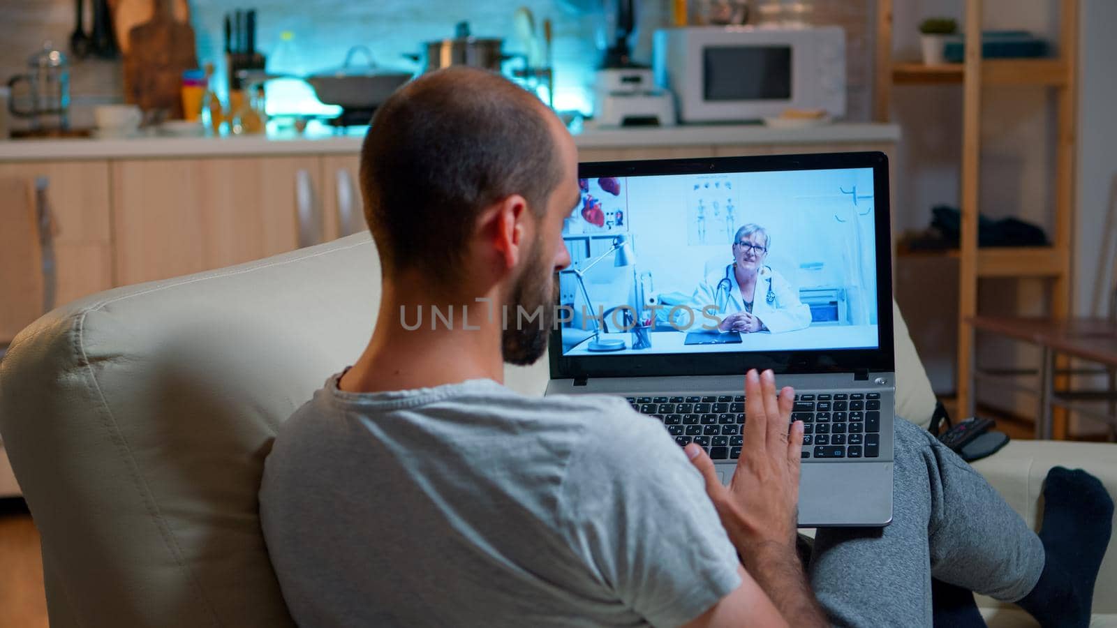 Physician doctor consulting online patient during coronavirus quarantine late at night in kitchen. Caucasian male sitting in front of laptop disscusing about illness symptoms and healthcare treatment