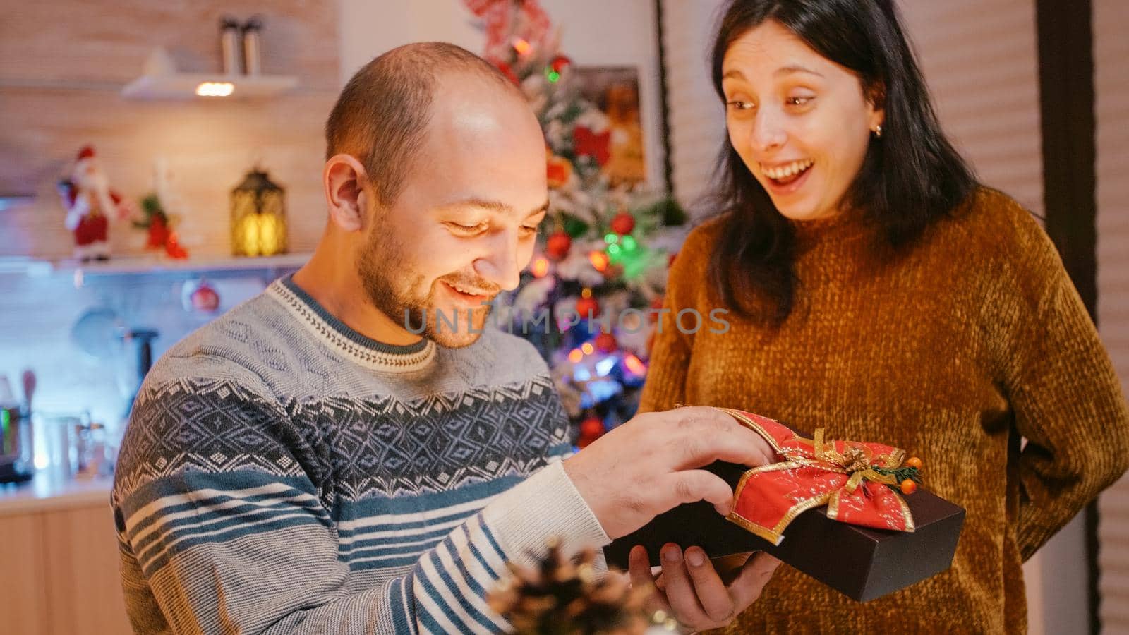Festive woman giving present box to man celebrating christmas eve. Couple kissing and hugging after opening gift with ribbon and seasonal ornaments for winter holiday celebration.