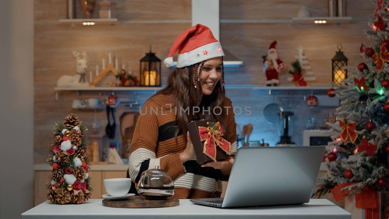 Festive woman using laptop for video call on christmas eve to exchange gifts online with friends and family. Person with santa hat showing present for seasonal spirit and celebration