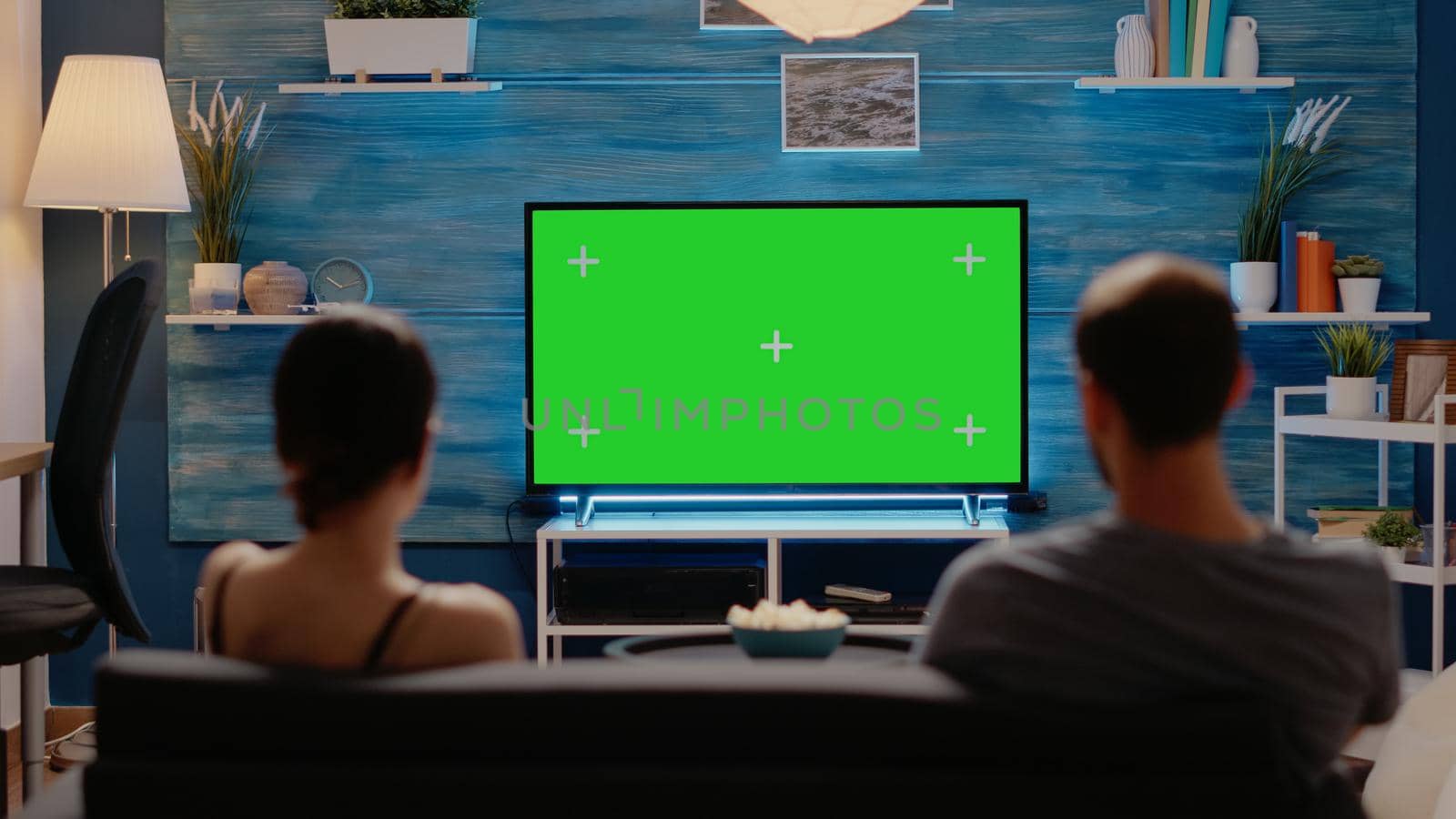 Caucasian man and woman looking at green screen on television display at home sitting in living room. Young couple with copy space on modern chroma key for isolated mockup template