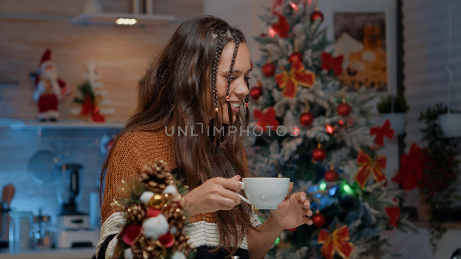 Caucasian woman holding cup of coffee while using video call in kitchen with christmas decorations. Young adult chatting with family while enjoying winter festivity wearing sweater