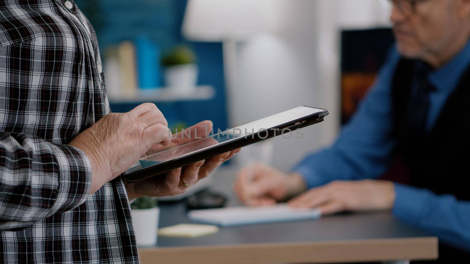 Close up of elderly woman employee texting, reading messages from tablet standing in home workspace while man working at laptop in background. Closeup of person hands holding notepad analysing graphs