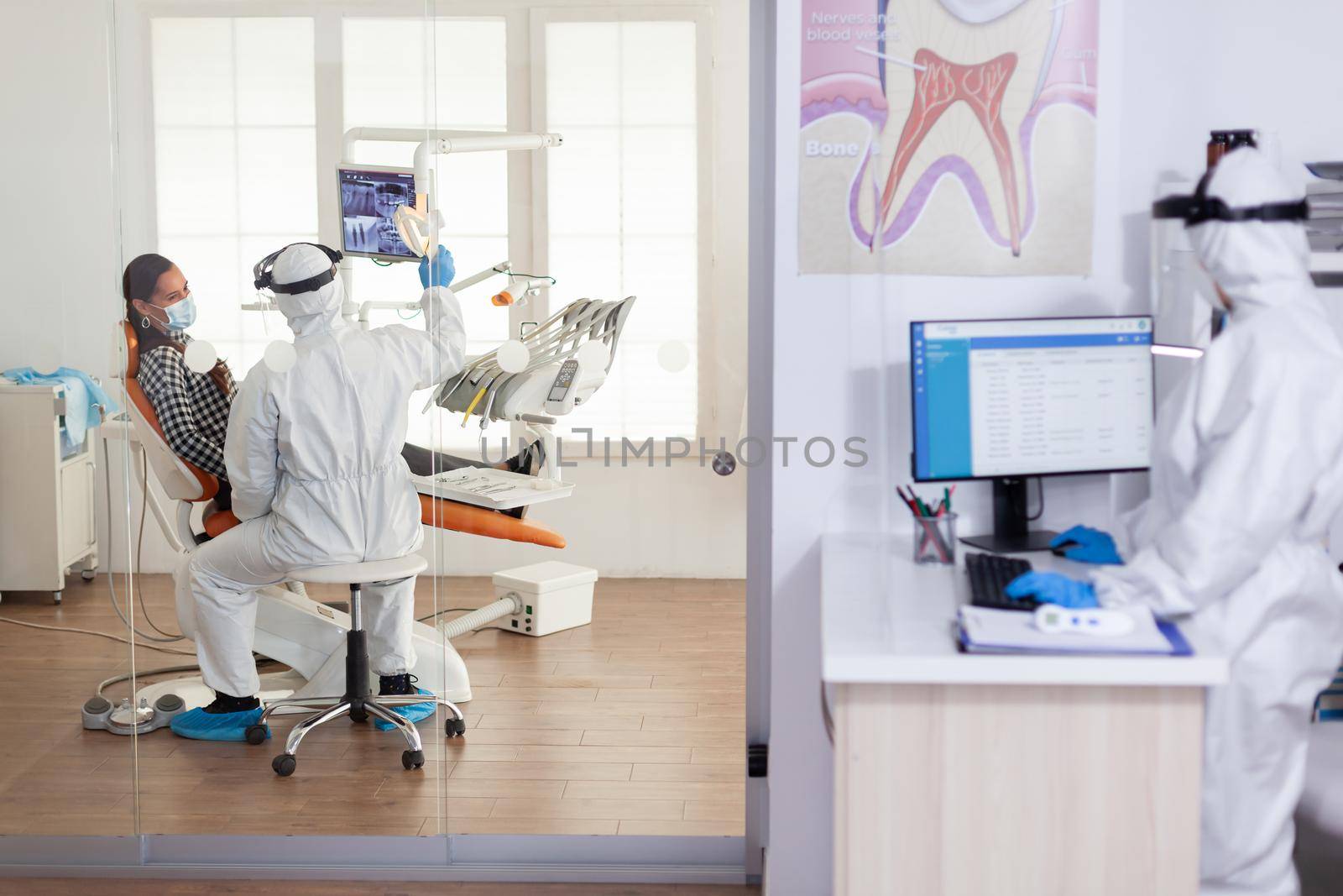 Stomatologist in protective suit asking for patient dental x-ray examining teeth problems during coronavirus pandemic in modern clinic. Medical team wearing coverall, face shield, mask and gloves.