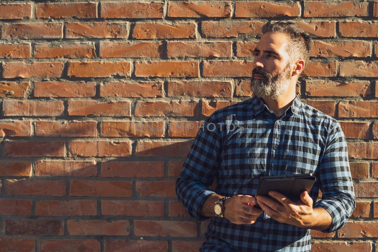 Portrait of modern businessman with beard thinking and using digital tablet while standing in front of brick wall outdoor.