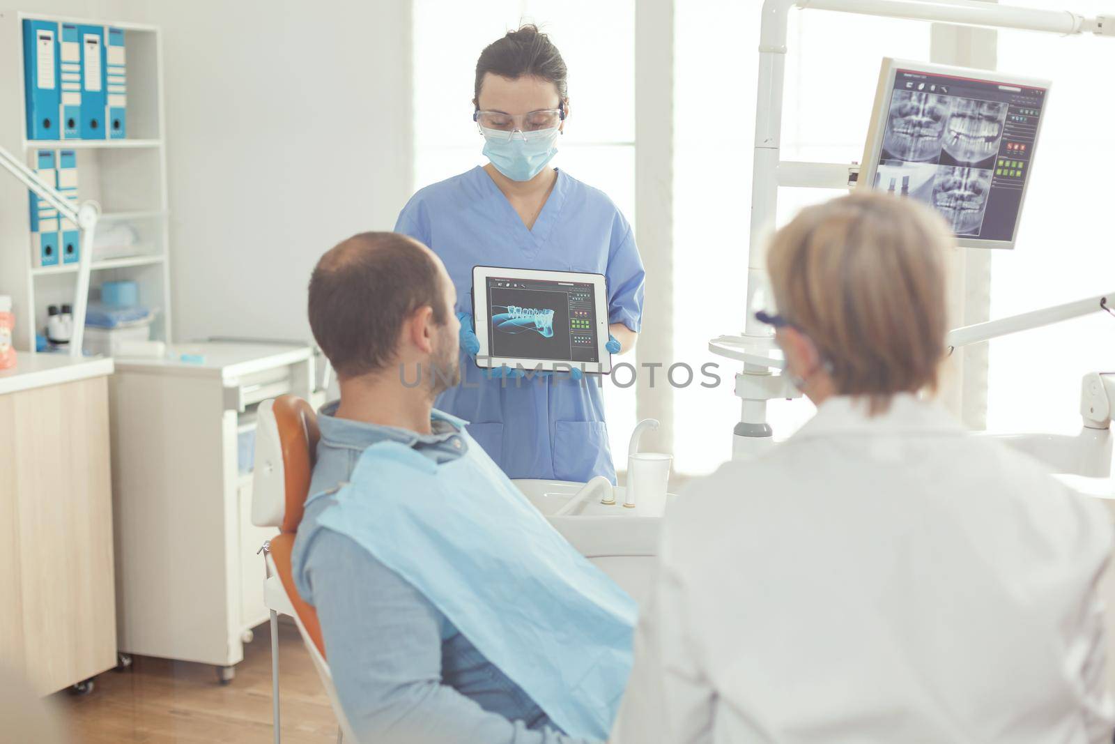 Stomatologist nurse showing tooth x-ray to sick patient explaining treatment using tablet working in stomatology hospital office. Senior doctor examining toothache while man sitting on dental chair