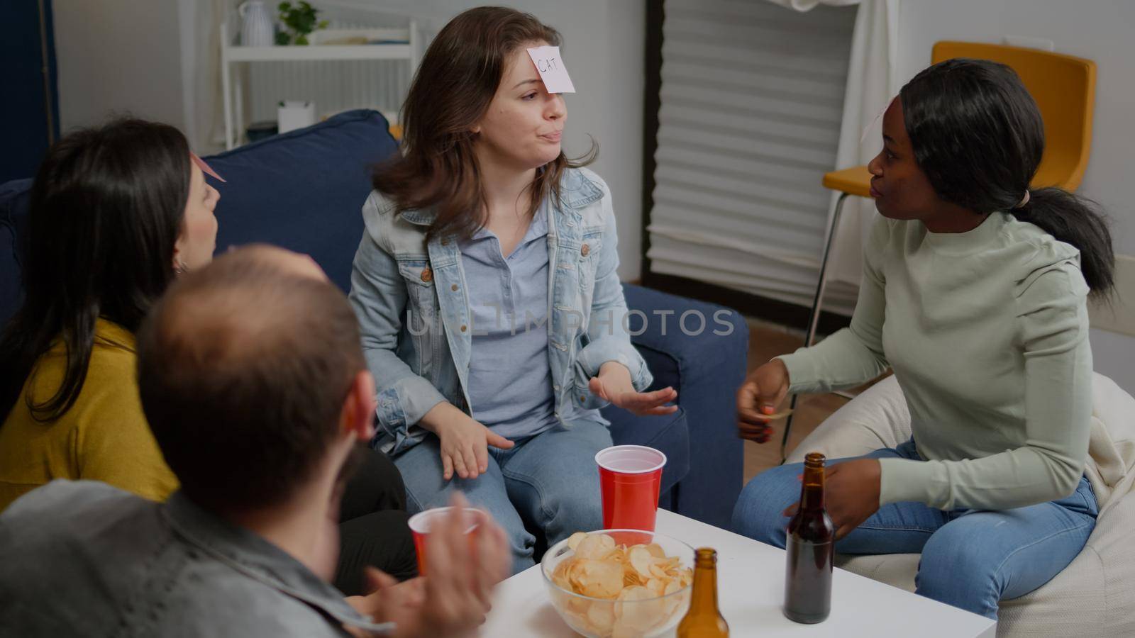 Mixed race friends gathering in living room playing guess who game using sticky notes. Mixed race people having fun, laughing together while sitting on sofa late at night during funny party.