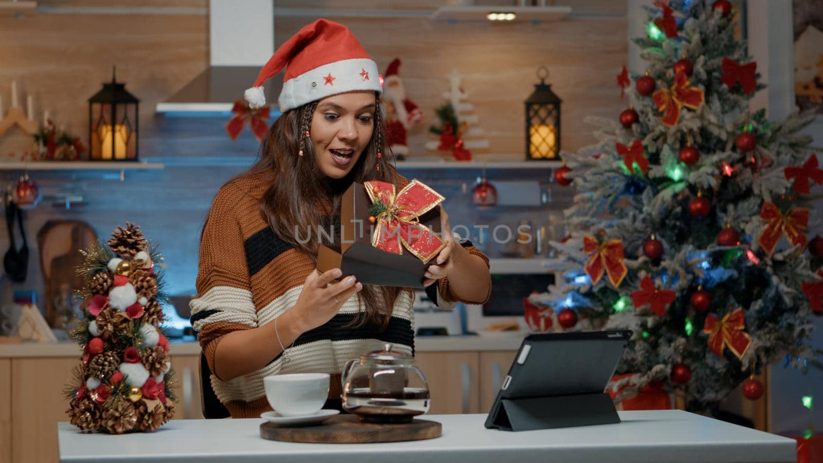 Smiling woman opening gift from friends on video call camera in festive home. Happy young adult using tablet at kitchen table receiving dream present for christmas eve celebration day