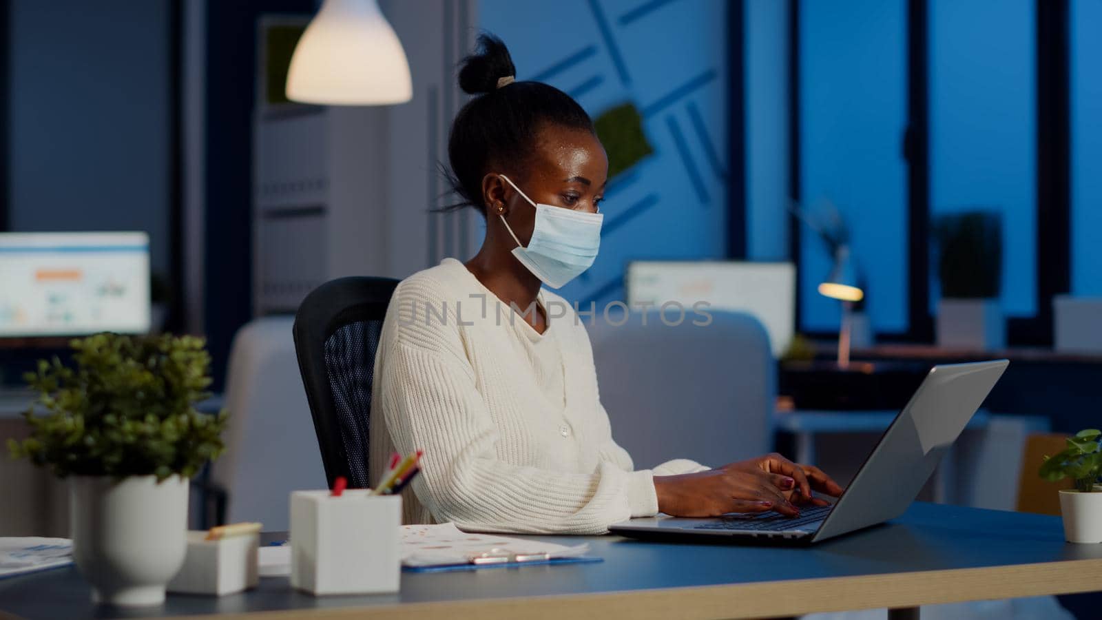 African employee with protection face mask working at laptop overtime in new normal business office to respect deadline of project, analysing documents sitting at desk overtime during global pandemic