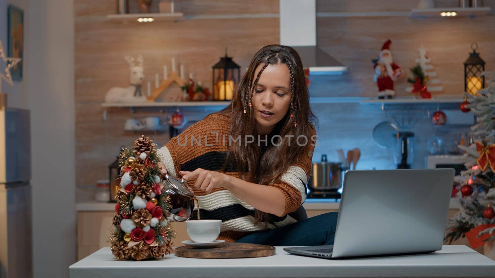Festive woman talking on video call using laptop in christmas decorated room. Young adult celebrating traditional holiday on internet comunication at home pouring cup of tea from kettle