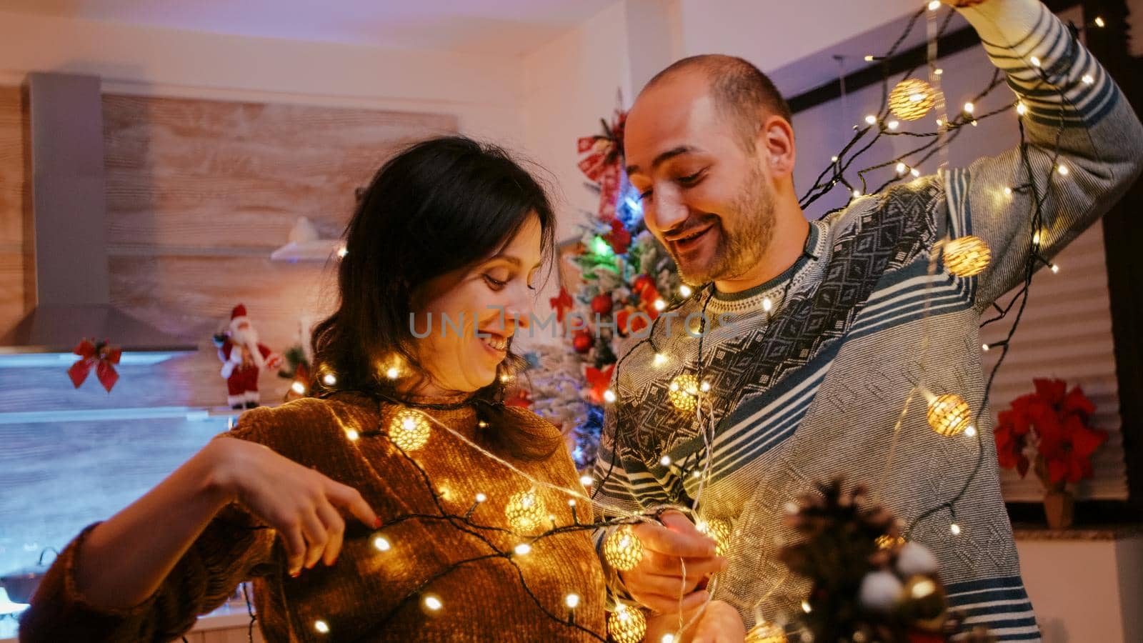 Man and woman tangled in garland of twinkle lights while decorating for christmas eve celebration. Couple laughing and untangling knot of string lights with illuminated bulbs. Cheerful people