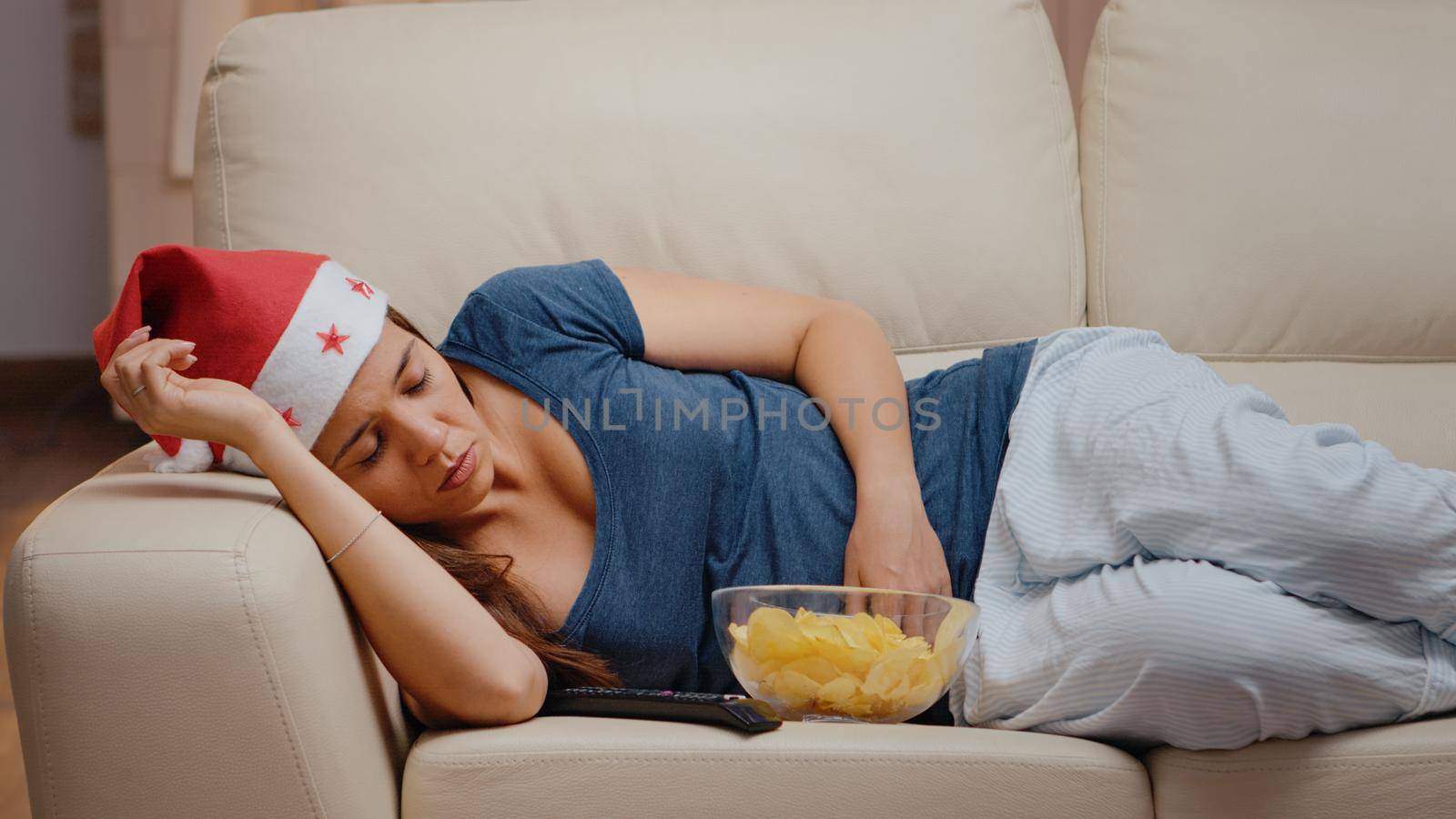 Close up of woman falling asleep while watching television on couch. Festive person wearing santa hat and feeling sleepy on christmas eve. Adult sleeping on sofa with bowl of chips