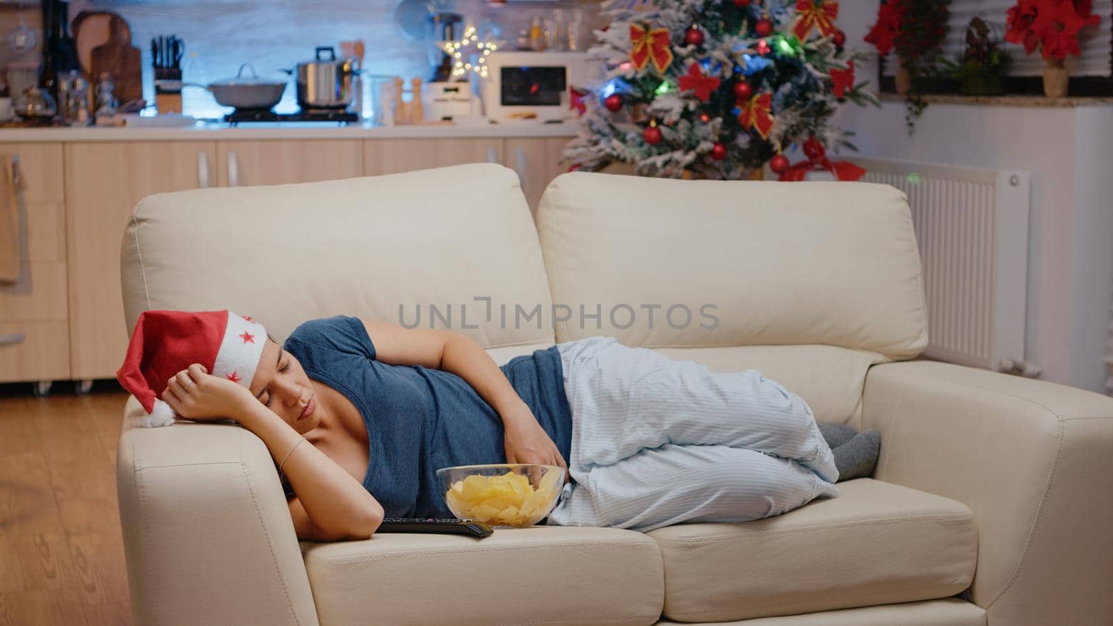 Sleepy woman wearing santa hat and resting on couch while looking at television. Person sleeping at TV, laying on sofa with snacks and remote control. Asleep adult on christmas eve