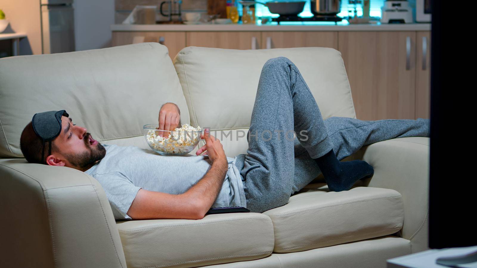 Caucasian man lying in sofa, eating popcron and watching TV late at night in the living room. Enjoying midnight snack, television entertainment, leisure lifestyle evening show