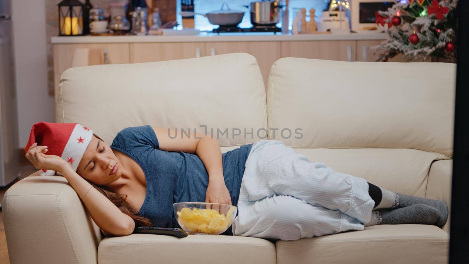 Sleepy woman with santa hat watching television on sofa. Festive adult falling asleep while looking at TV screen with TV remote control and bowl of chips. Person sleeping on christmas eve.