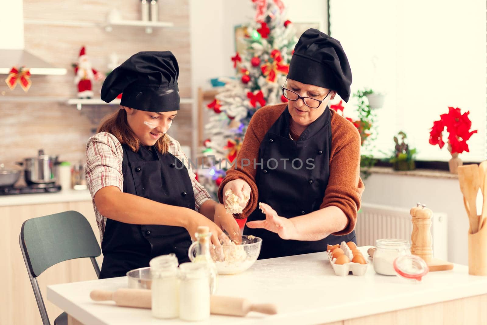 Kid wearing apron mixing dough on christmas day with grandmother. Happy cheerful joyfull teenage girl wearing apron and bonette helping senior woman preparing sweet cookies to celebrate winter holidays.