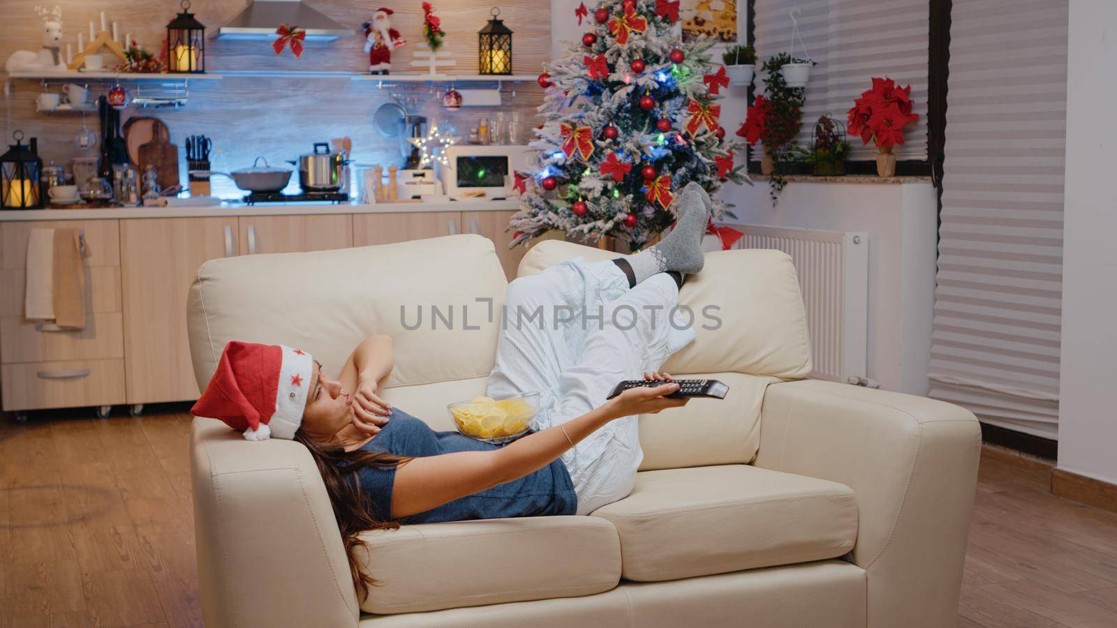 Bored adult switching channels with TV remote control while laying on couch. Woman wearing santa hat and watching television, eating chips from bowl on christmas eve festivity.