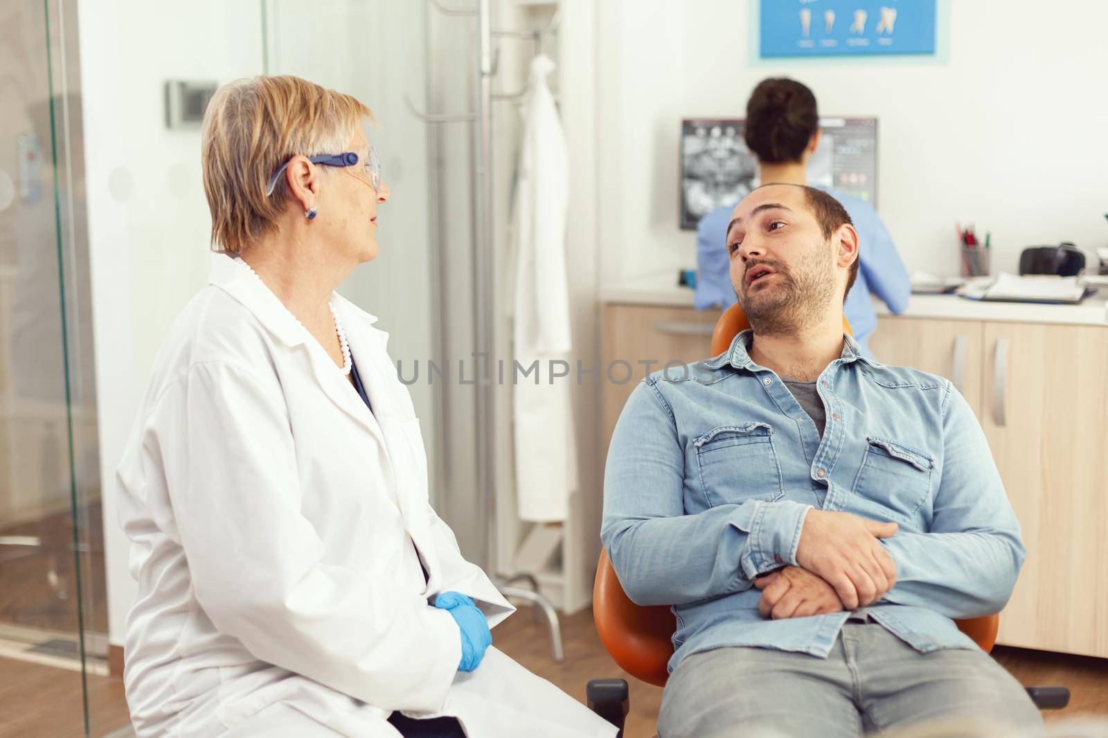 Senior doctor stomatologist discussing with patient before examining oral health while sitting on dental chiar in hospital stomatology office. In background nurse checking tooth radiography