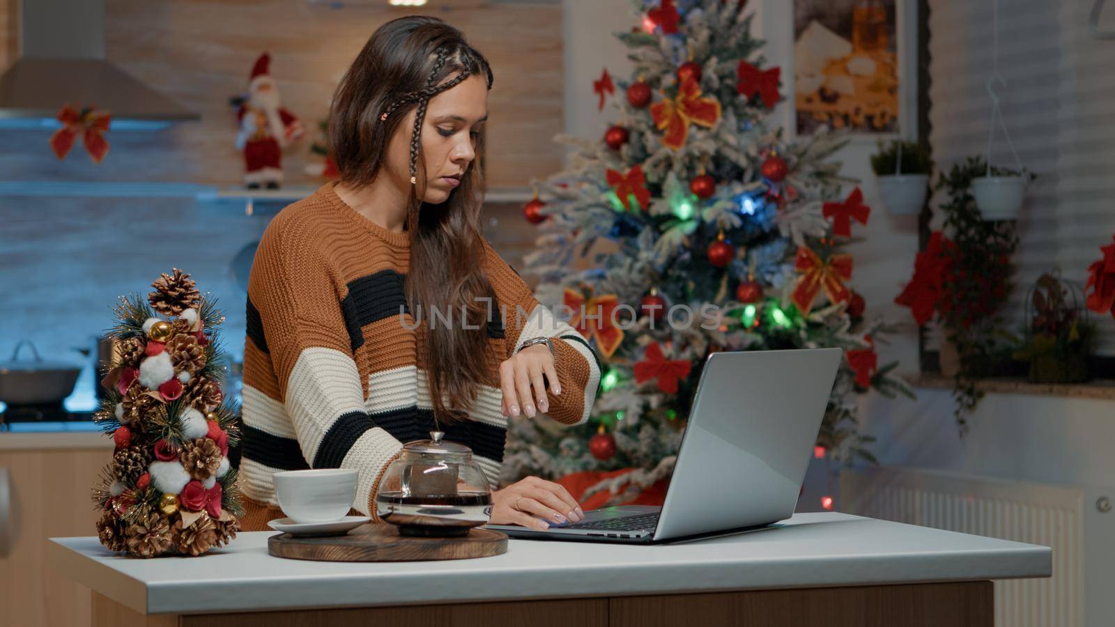 Sad woman waiting for friends to start christmas celebration in kitchen with decorations, ornaments and tree. Adult using laptop before preparing for guests at holiday dinner party