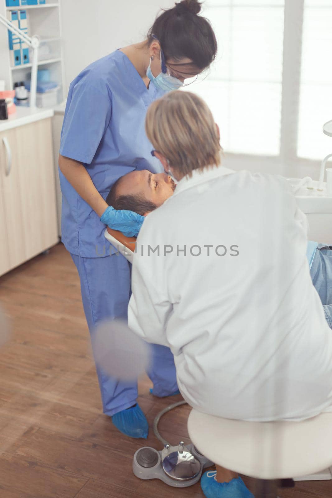 Sick patient sitting on stomatological chair with open mouth during dental examination. Senior woman stomatologist and medical nurse with masks holding sterilized tools checking teeth health