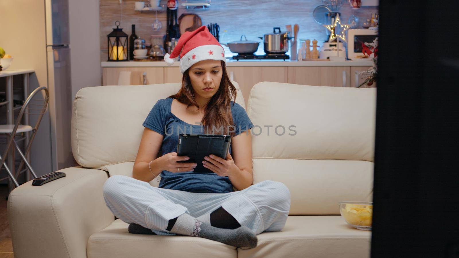 Adult using digital tablet and watching movie on television for christmas eve celebration. Woman looking at gadget and TV screen alone on seasonal holiday festivity. Person wearing hat