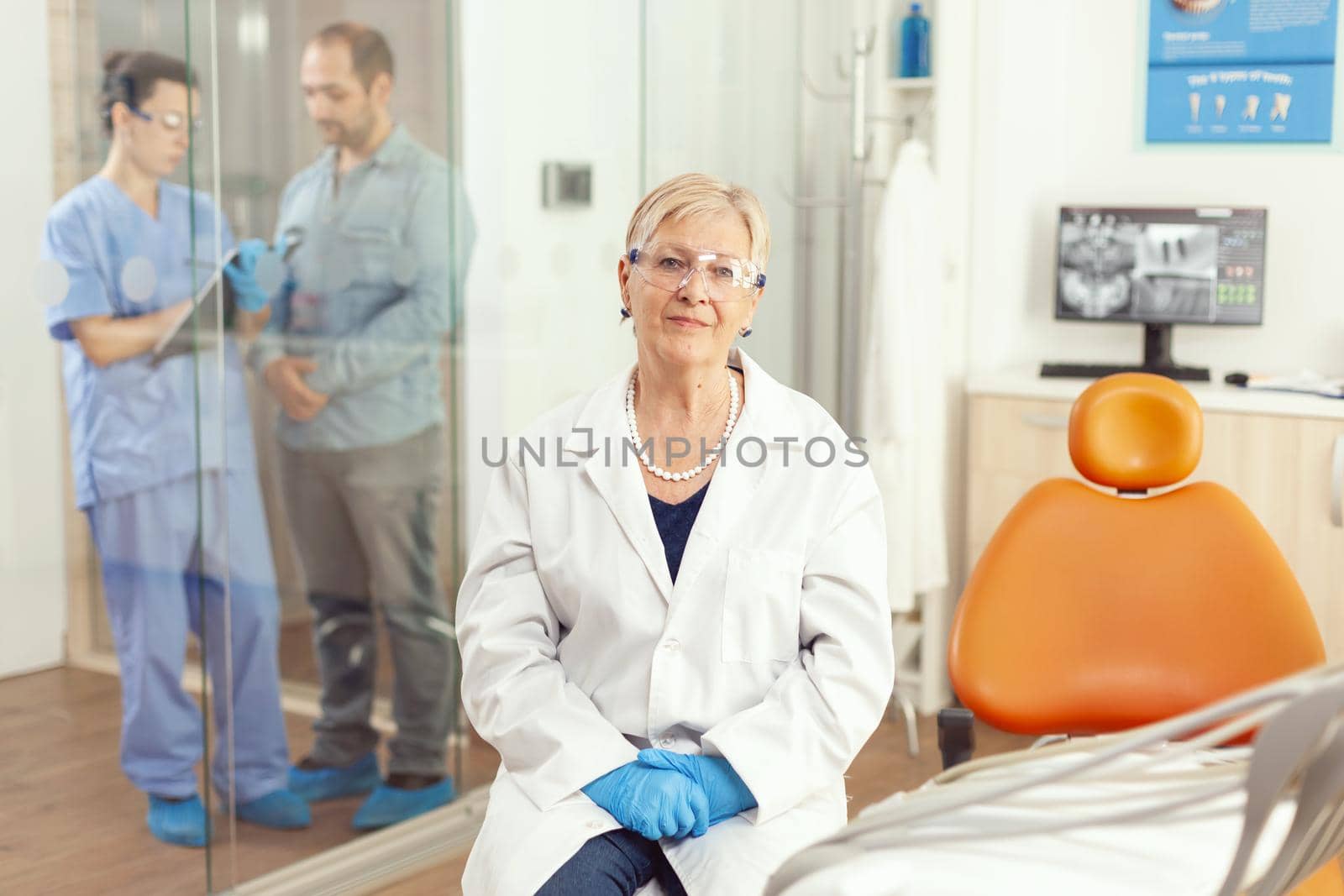 Senior stomatological woman looking into camera while sitting on dental chair in hospital stomatology room. Medical nurse discussing with patient during writing healdcare treatment