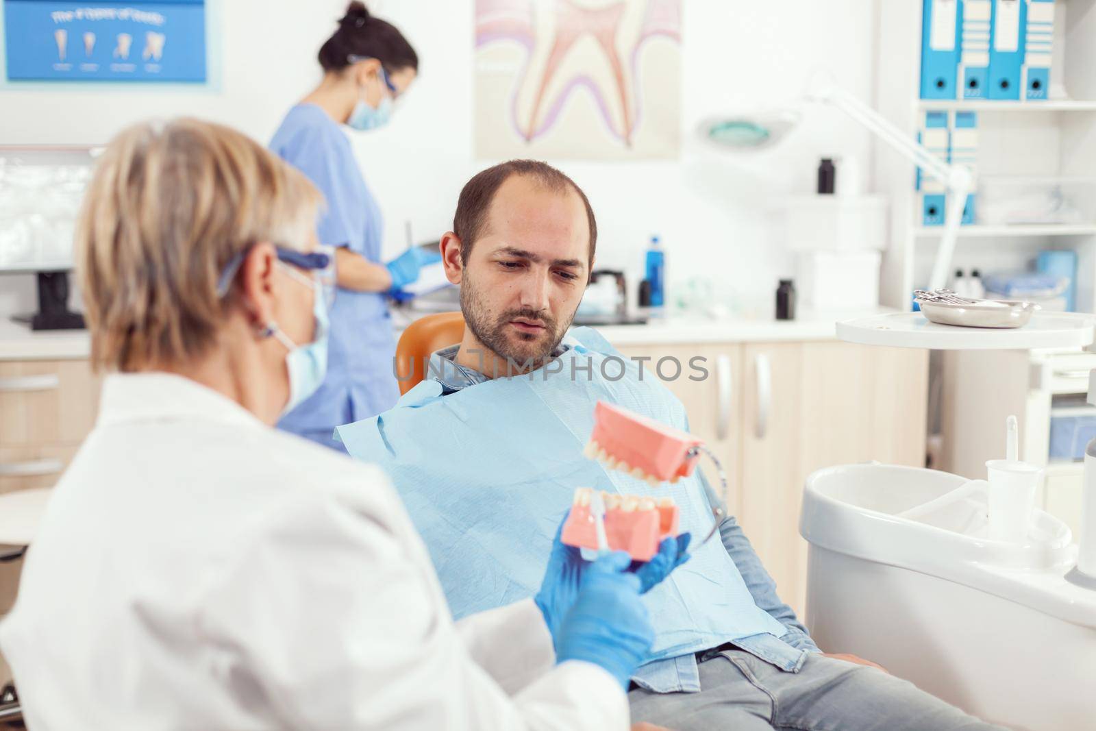 Stomatologist senior woman holding medical tooth skelaton explaining dental hygiene to man patient for prevent tooth surgery while sitting on stomatology chair. Doctor examining sick man