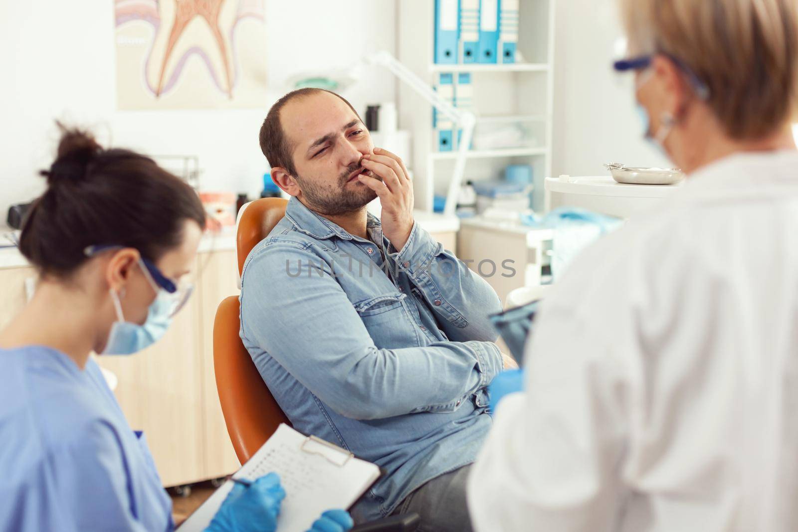 Sick patient with toothache speaking with stomatologist showing affected mass. Man explaining dental problem to doctor while nurse writing stomatological treatment in modern private clinic