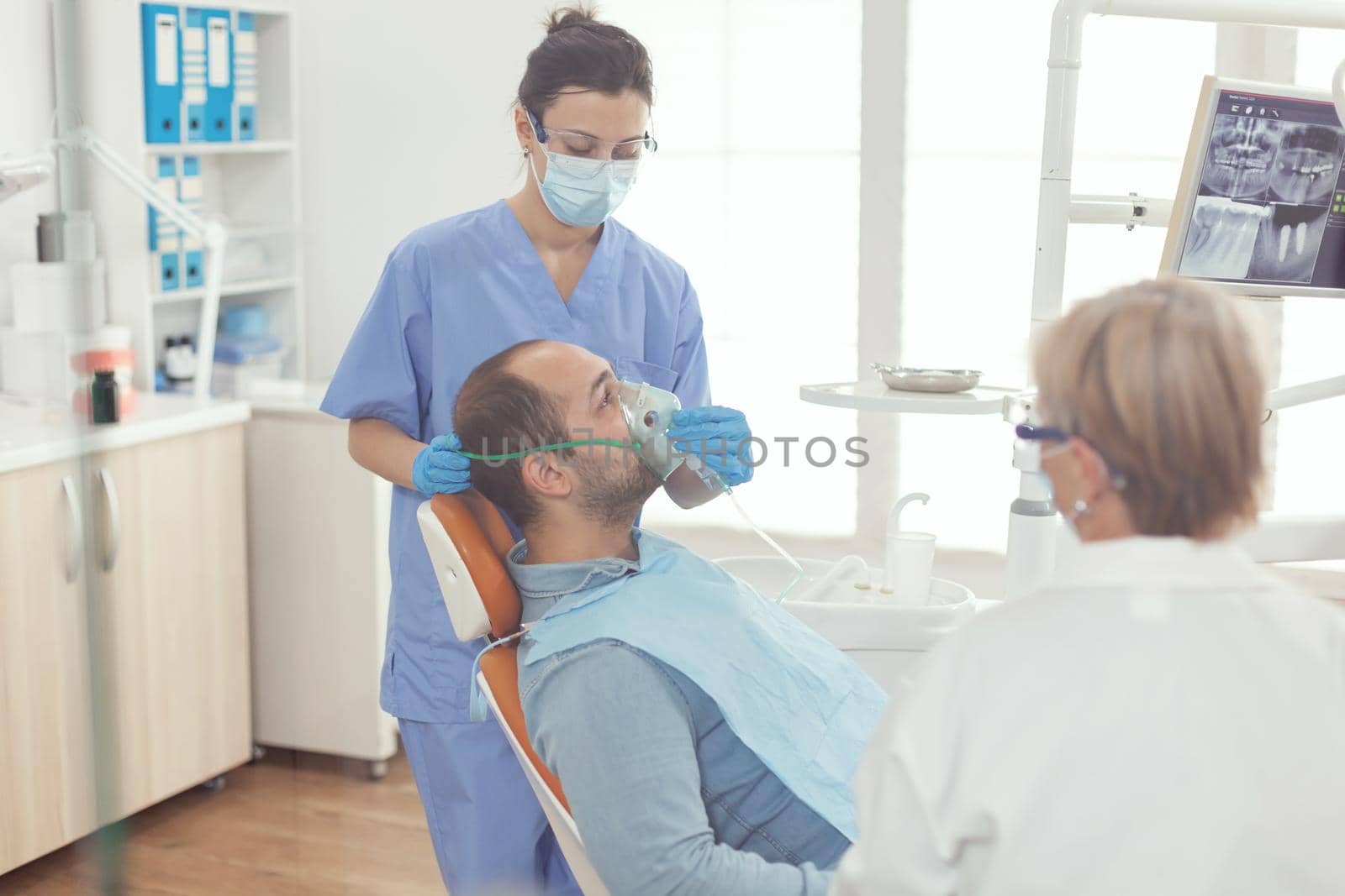Medical nurse putting oxigen mask preparing sick man patient for stomatology surgery while sitting on dental chair in orthodontic hospital room. Dentist doctor examining toohache