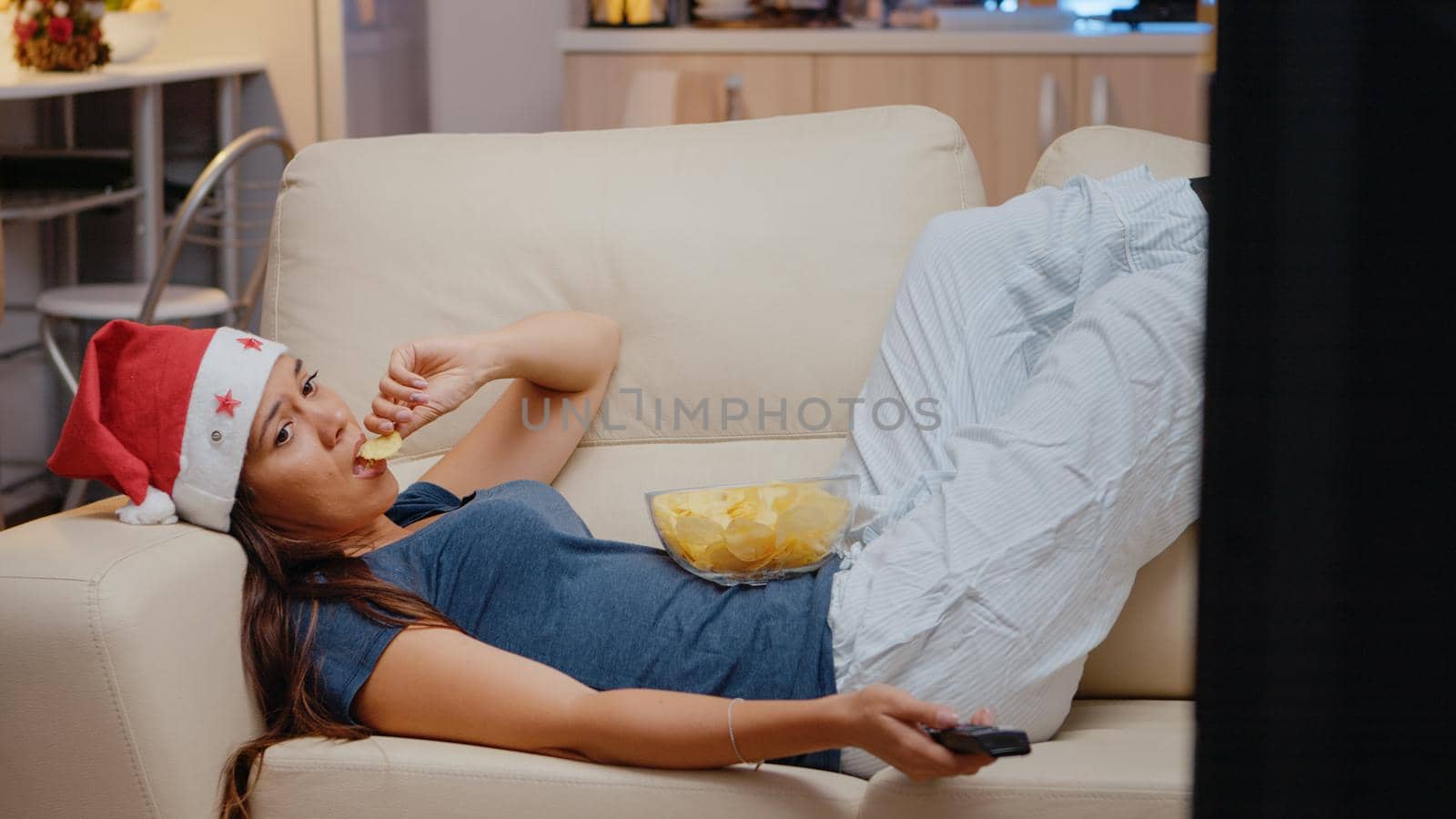 Sad woman laying on couch and watching television with TV remote control on christmas eve. Bored adult wearing santa hat and looking at screen while eating chips from bowl on holiday