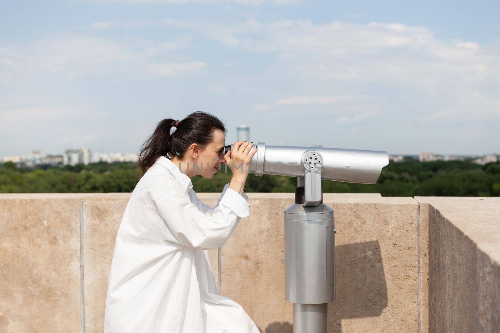 Young tourist woman standing on building rooftop looking through telescope by DCStudio