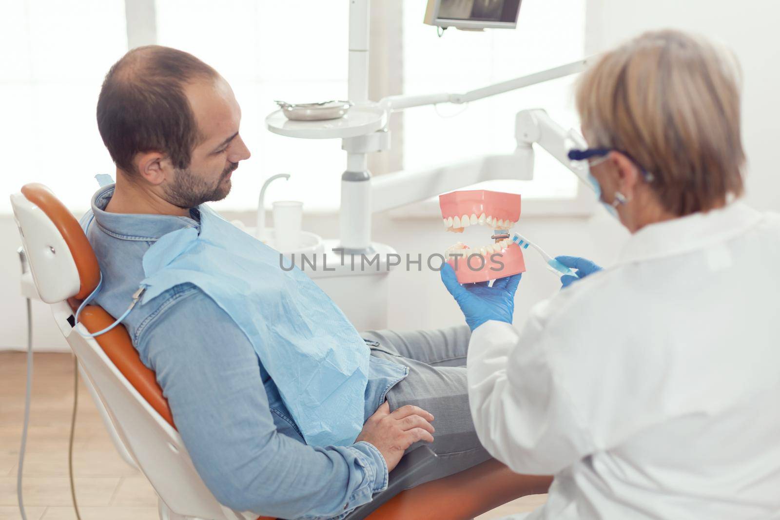 Senior woman dentist explaining tooth surgery using plaster model of mandible. Stomatologist holding mock-up of skeleton of teeth during stomatology appointment working in medical office