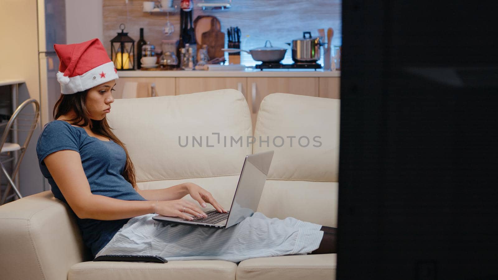 Festive woman working on laptop while watching television on christmas eve holiday. Adult using gadget and looking at TV, sitting on couch with santa hat and decorations for festivity.