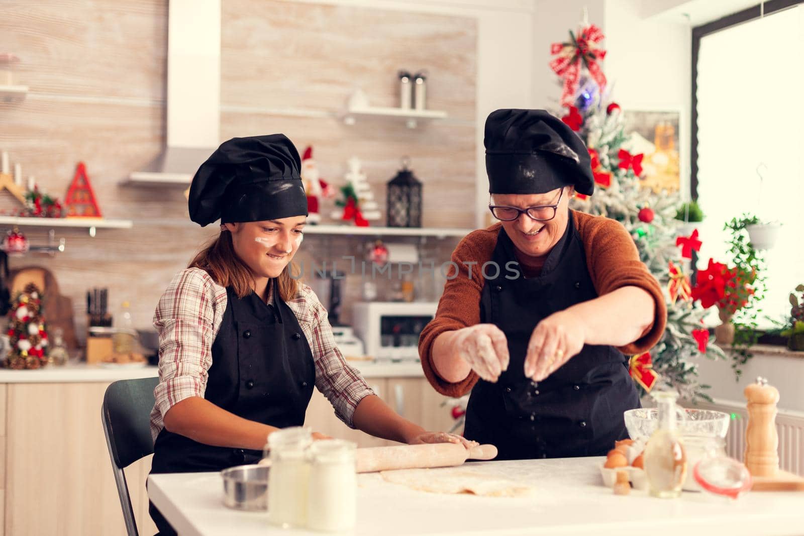 Grandmother in apron during christmas day baking making desert with child. Happy cheerful joyfull teenage girl helping senior woman preparing sweet cookies to celebrate winter holidays.
