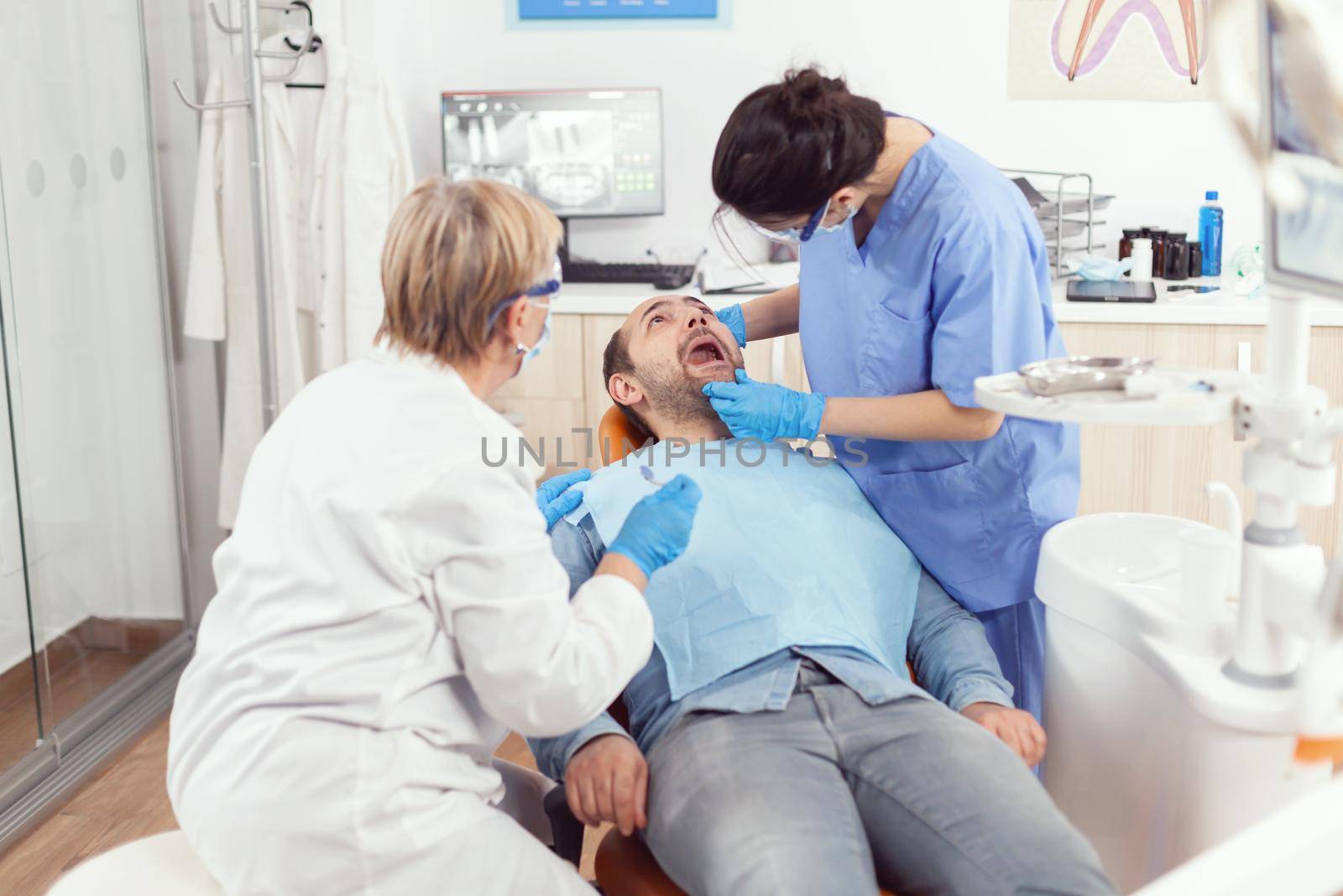 Sick man sitting on dental chair with open mouth while medical nurse with face mask and gloves analyzing teeth health during stomatology consultation. Hospital team working in stomatological clinic