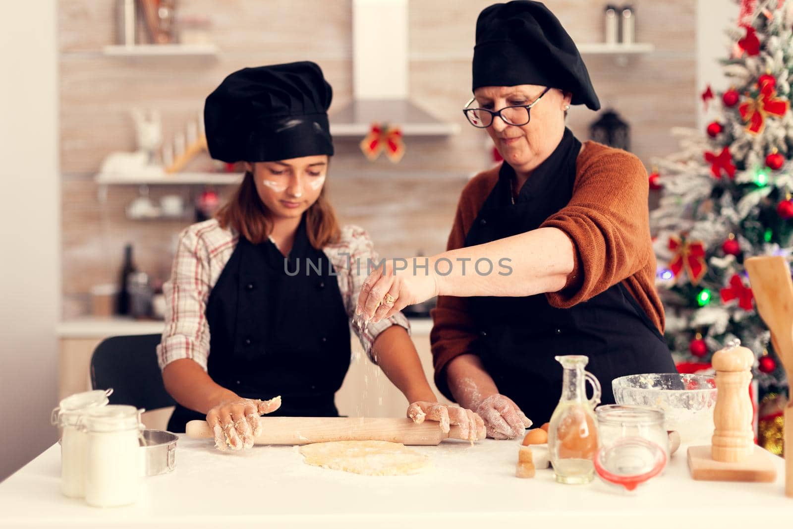 Grandchild wearing apron on christmas day and grandmother spreading flour over dough. Happy cheerful joyfull teenage girl helping senior woman preparing sweet cookies to celebrate winter holidays.