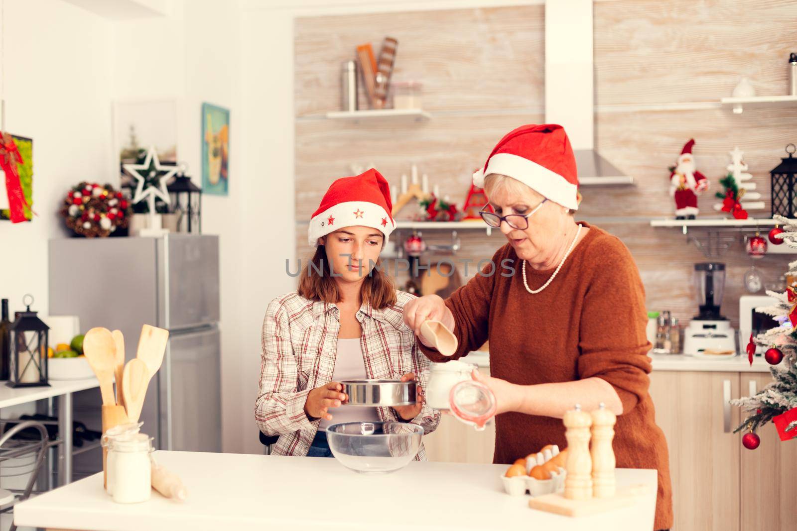 Niece on christmas day making cookies with grandmother in decorated kitchen. Happy cheerful joyfull teenage girl helping senior woman preparing sweet biscuits to celebrate winter holidays wearing santa hat.