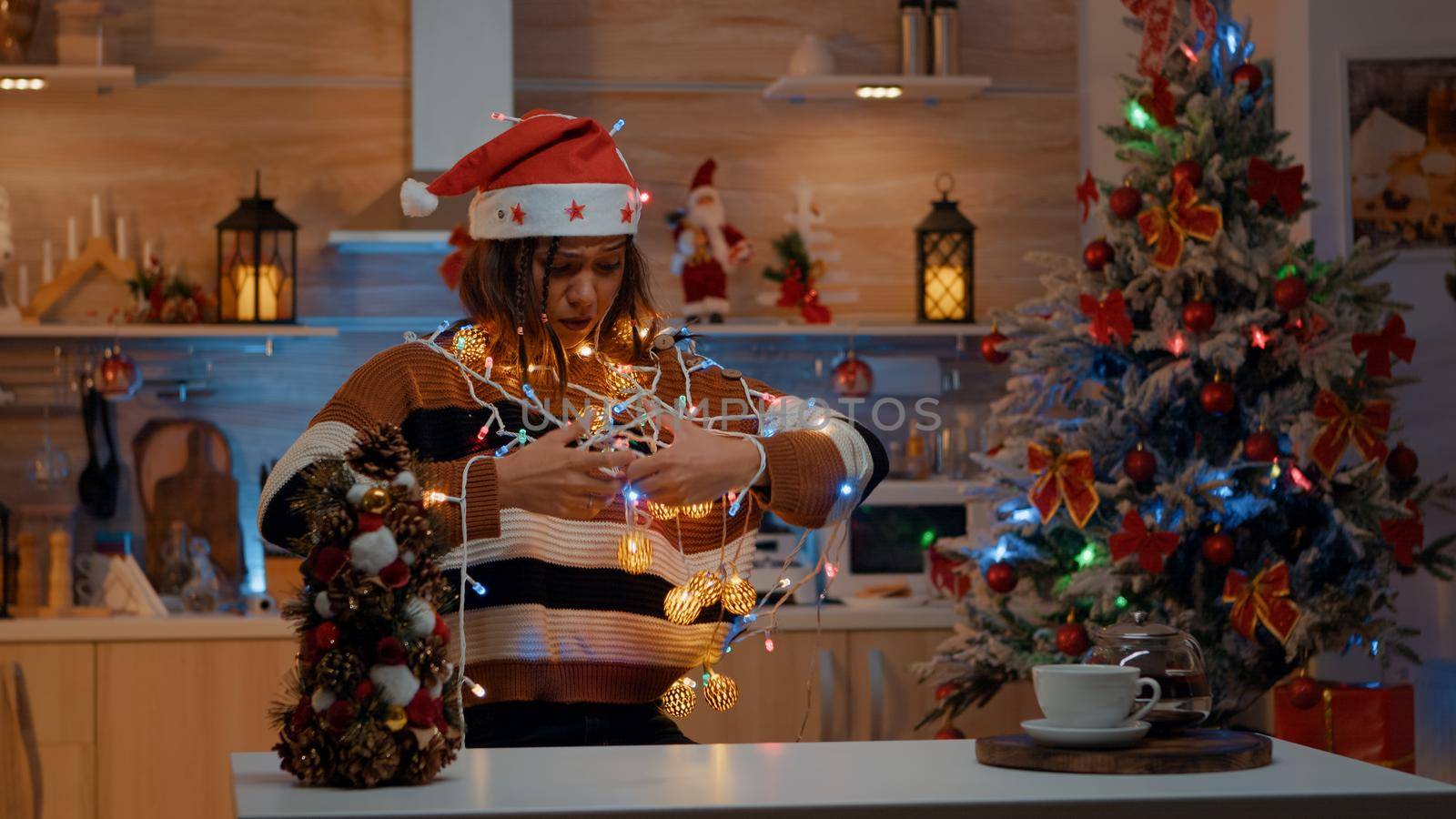 Young person holding illuminated string garland for christmas decorations in kitchen at home. Woman getting tangled in shiny decor lights while preparing for holiday celebration party