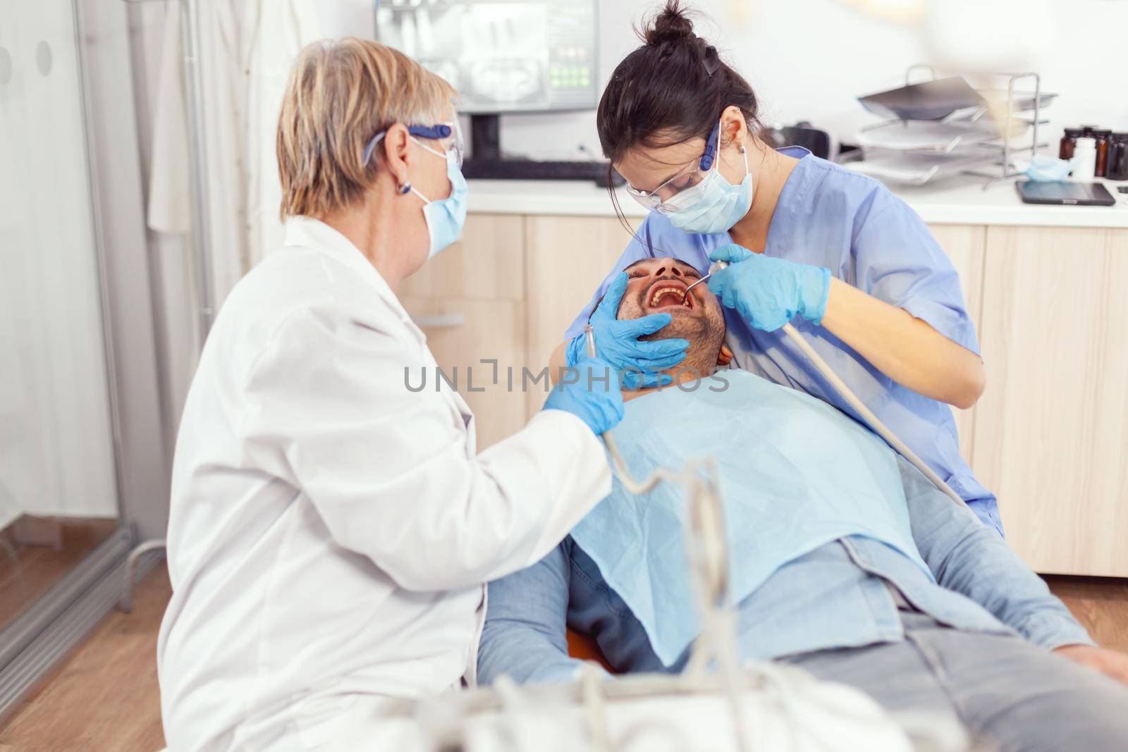 Sick man sitting on medical chair with open mouth while medical nurse with face mask and gloves cleaning teeth after dental surgery. Hospital team working in stomatological clinic