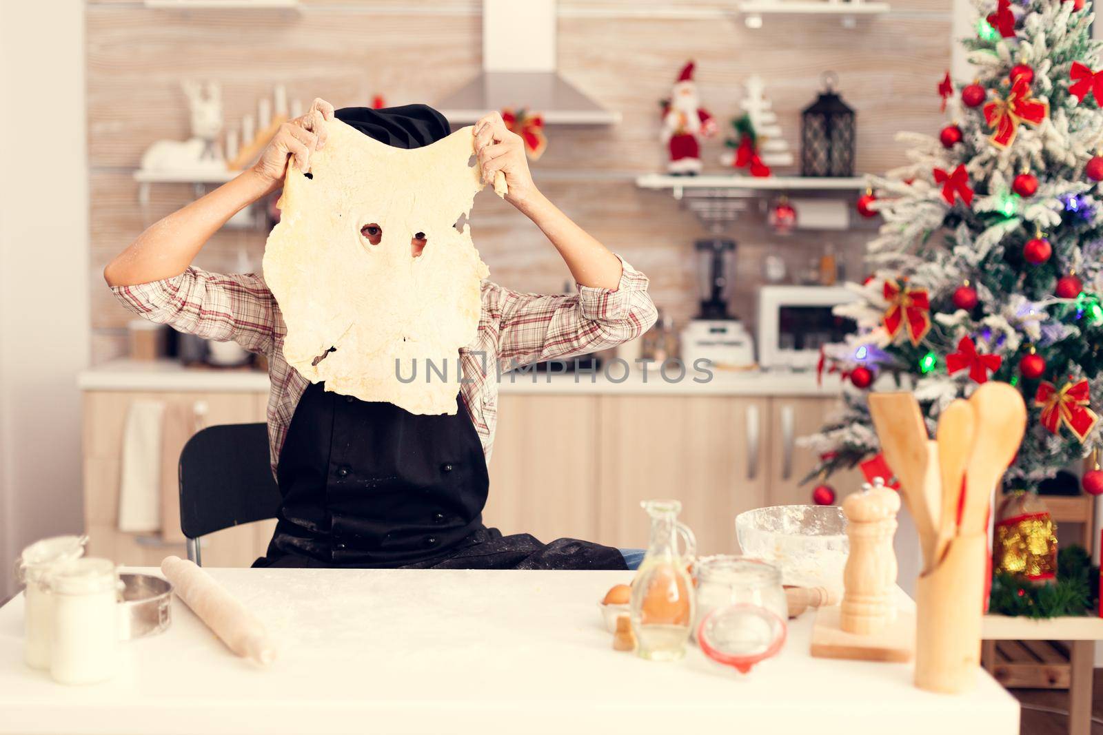 Cute kid doing traditional dessert on christmas day wearing apron and bonette. Cheerful happy cute girl while prepearing delicious cookies for christmas celebration in kitchen with christmas tree in the background.