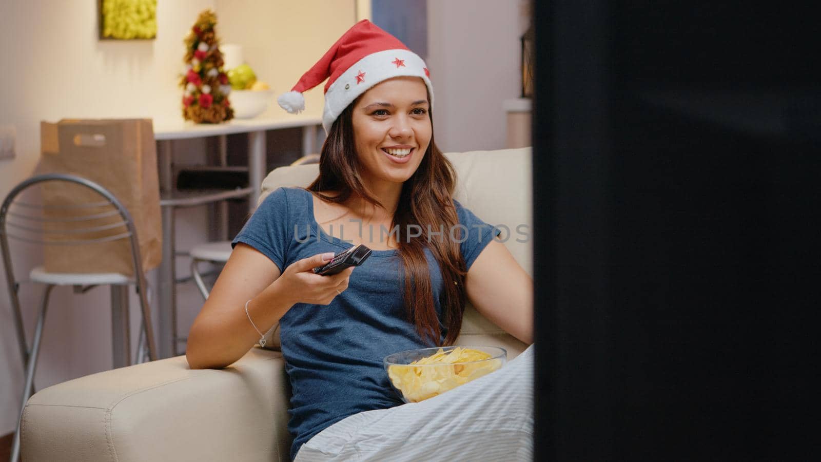 Cheerful woman watching television and eating chips on christmas eve festivity. Person wearing santa hat and holding TV remote control while looking at comedy movie and laughing