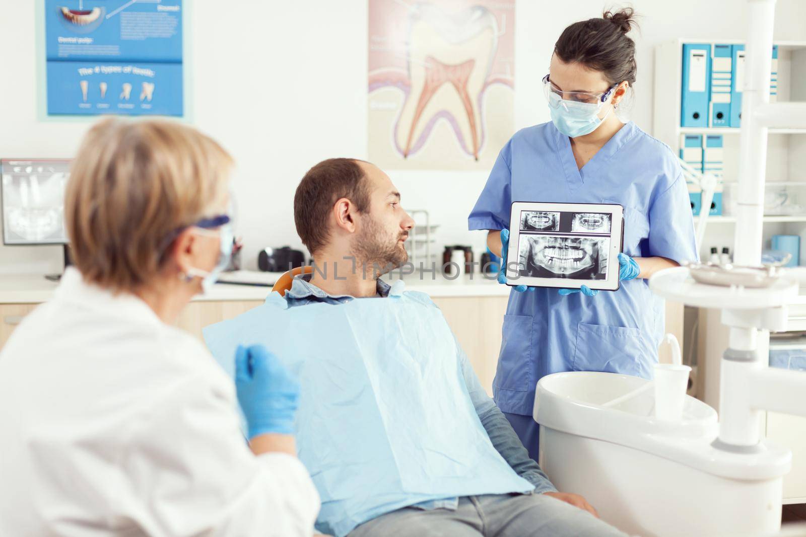 Sick man sitting on dental chaiar looking at radiography on digital tablet discussing with senior doctor stomatologist about toothache treatment. Medical stomatology team working in dentistry office