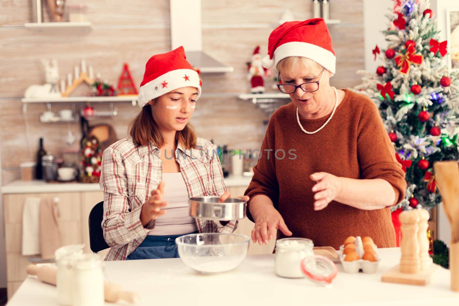 Child on christmas day with xmas tree in the background baking traditional cookies. Happy cheerful joyfull teenage girl helping senior woman preparing sweet biscuits to celebrate winter holidays wearing santa hat.