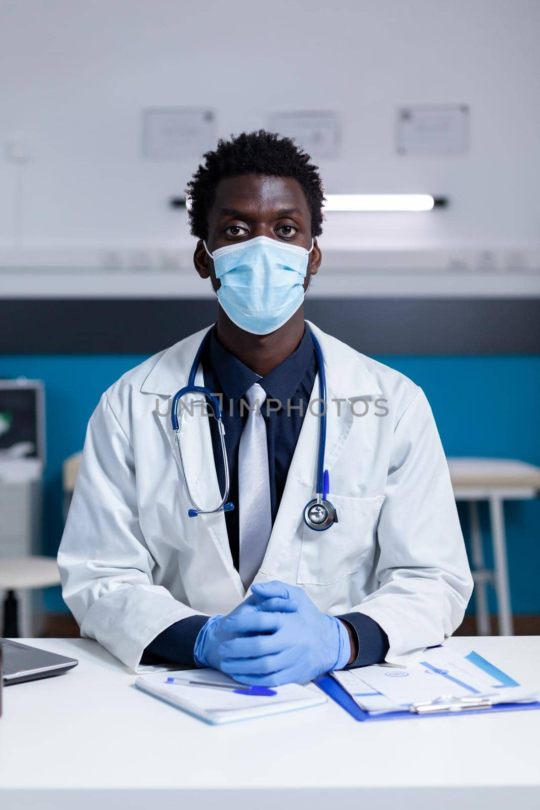Doctor of african american ethnicity sitting at desk with face mask and stethoscope looking at camera in medical cabinet. Black medic wearing gloves and coat at healthcare clinic