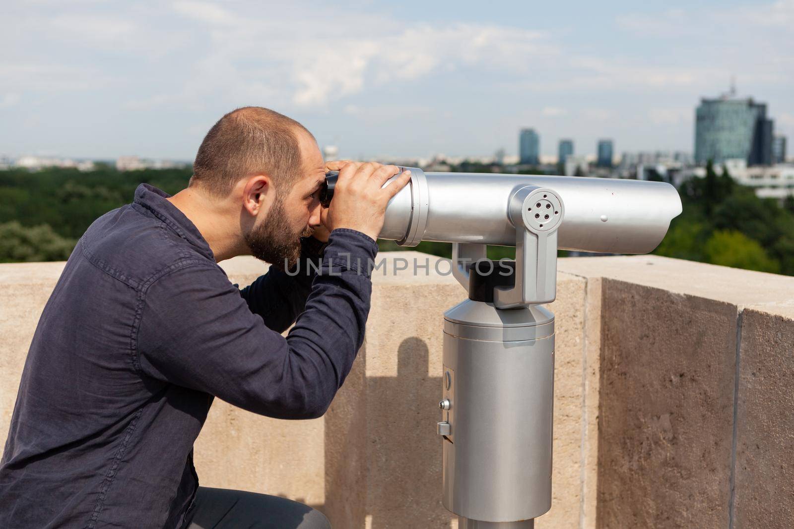 Man wathching through binoculars telescope standing on tower rooftop at observation point. Tourist enjoying metropolitan urban city landscape. Travel concept. Panoramic view