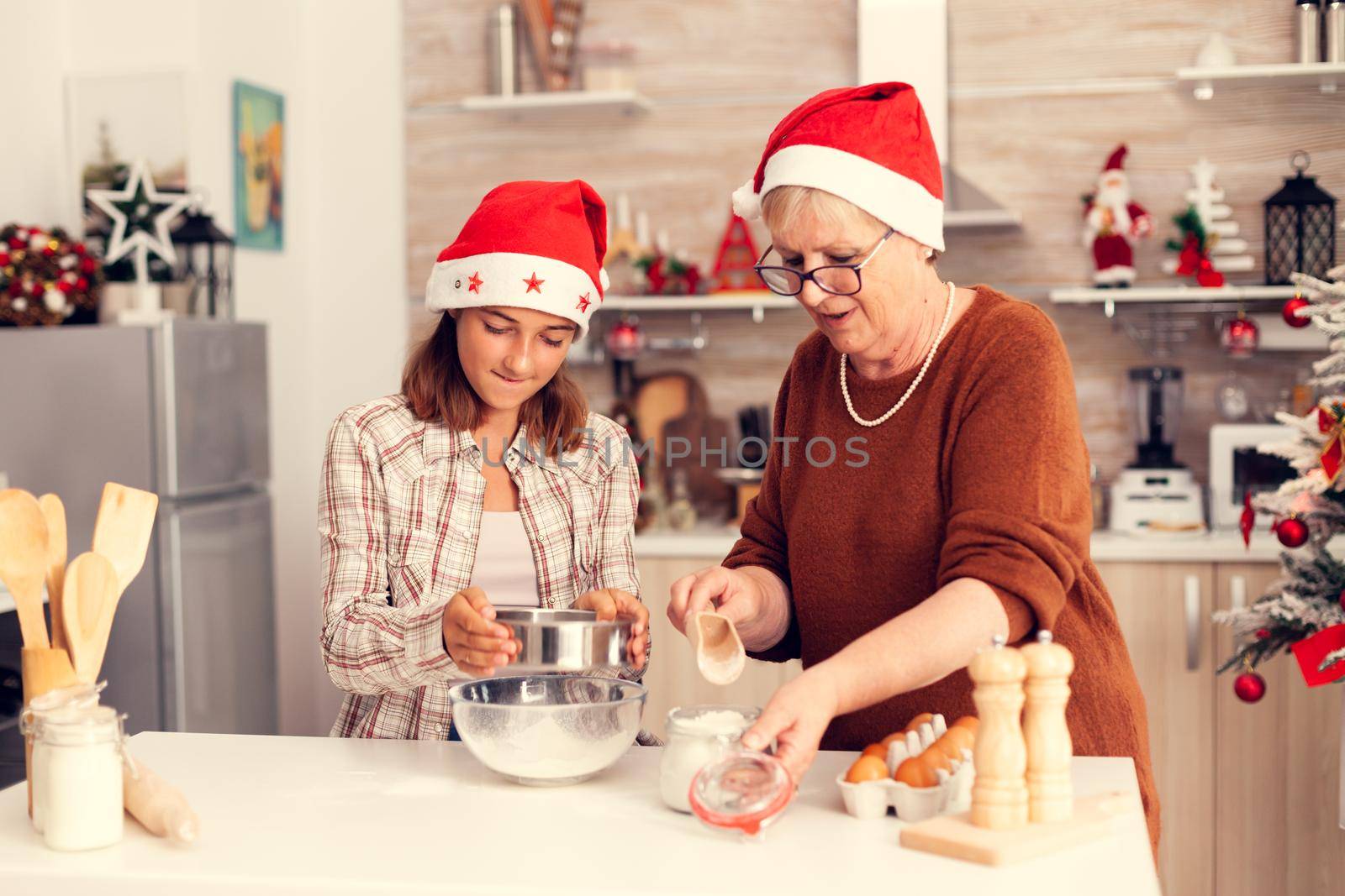 Grandchild on christmas day having fun in kitchen with gandmother praparing tasty cookies. Happy cheerful joyfull teenage girl helping senior woman preparing sweet biscuits to celebrate winter holidays wearing santa hat.