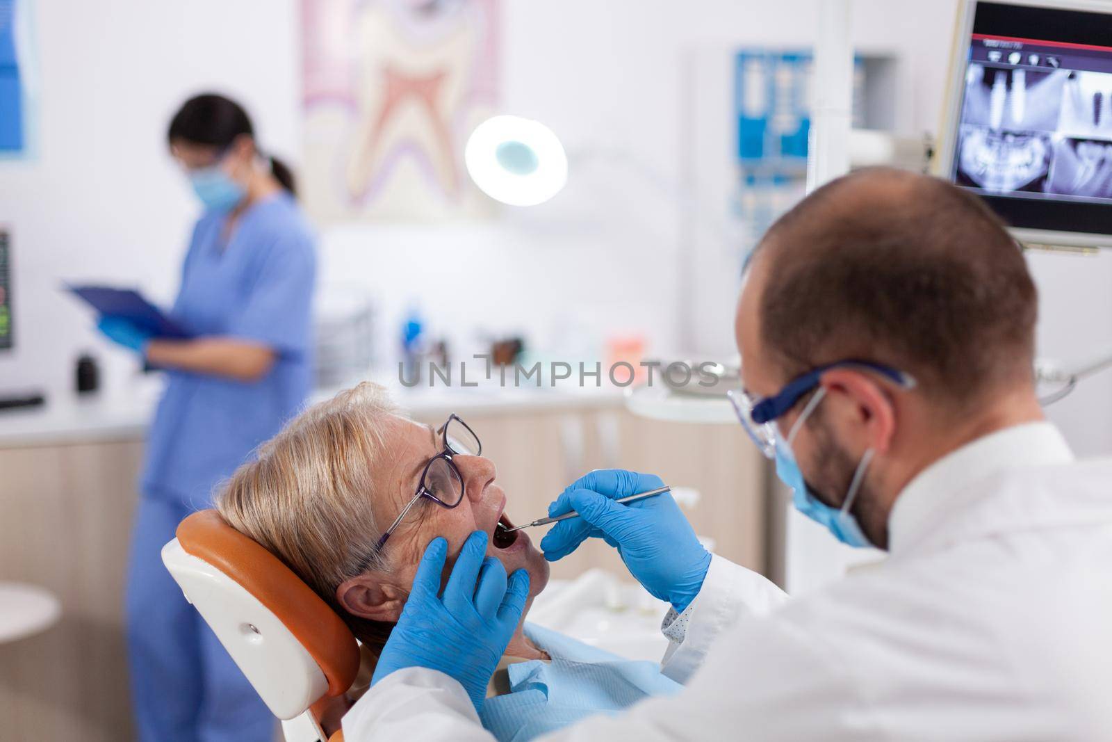 Stomatolog wearing face mask during consultation of senior woman using angled mirror. Elderly patient during medical examination with dentist in dental office with orange equipment.