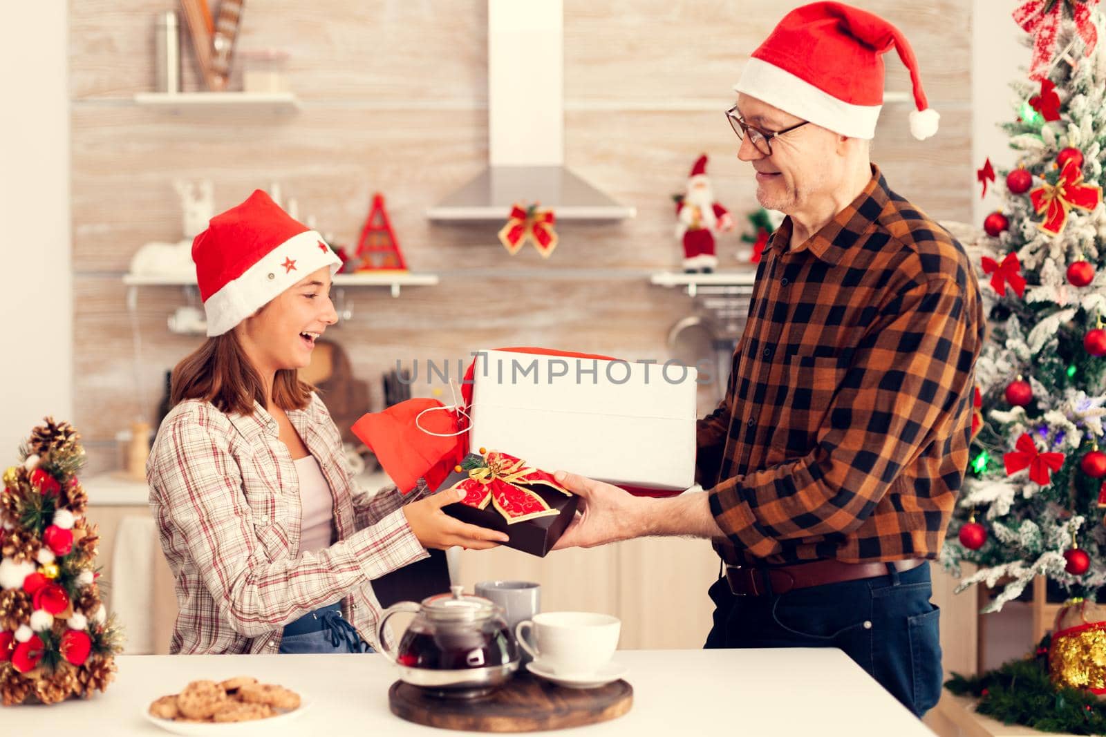 Happy grandchild exchanging presents with grandfather celebrating christmas. Senior man wearing santa hat surprising granddaughter with winter holiday gifts in home kitchen with xmas tree in the background.