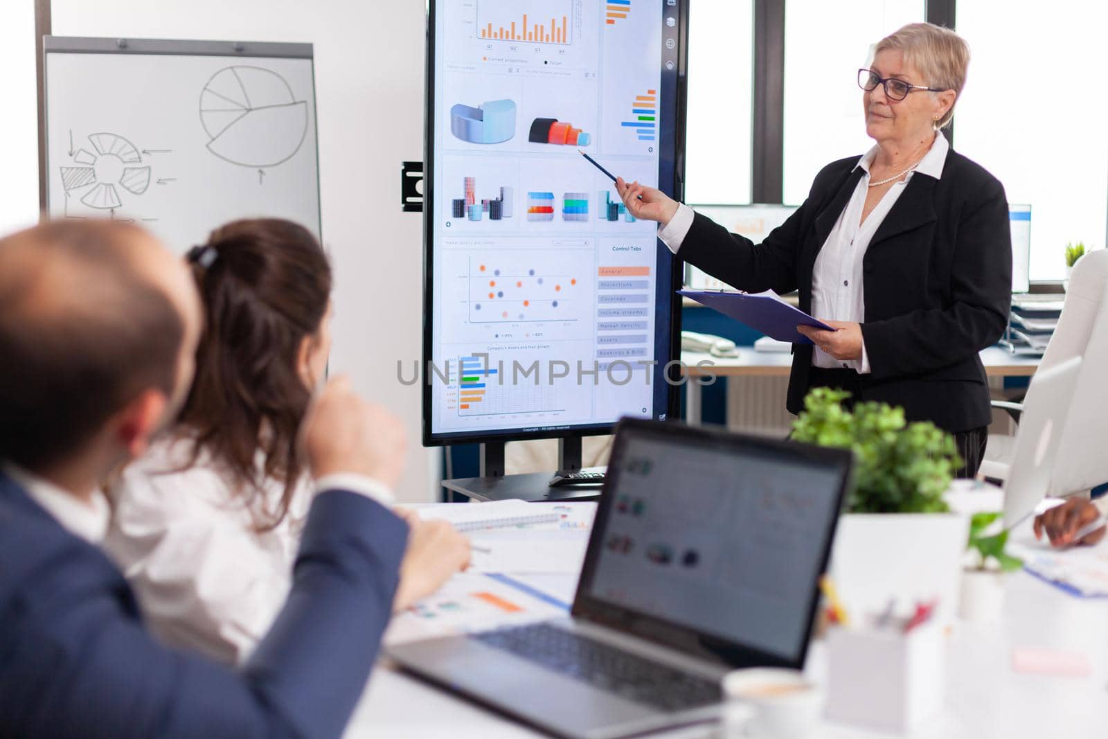 Elderly project manager pointing at desktop presenting statistical data, briefing diverse group of employees. Multiethnical businesspeople working in professional startup financial office during conference meeting