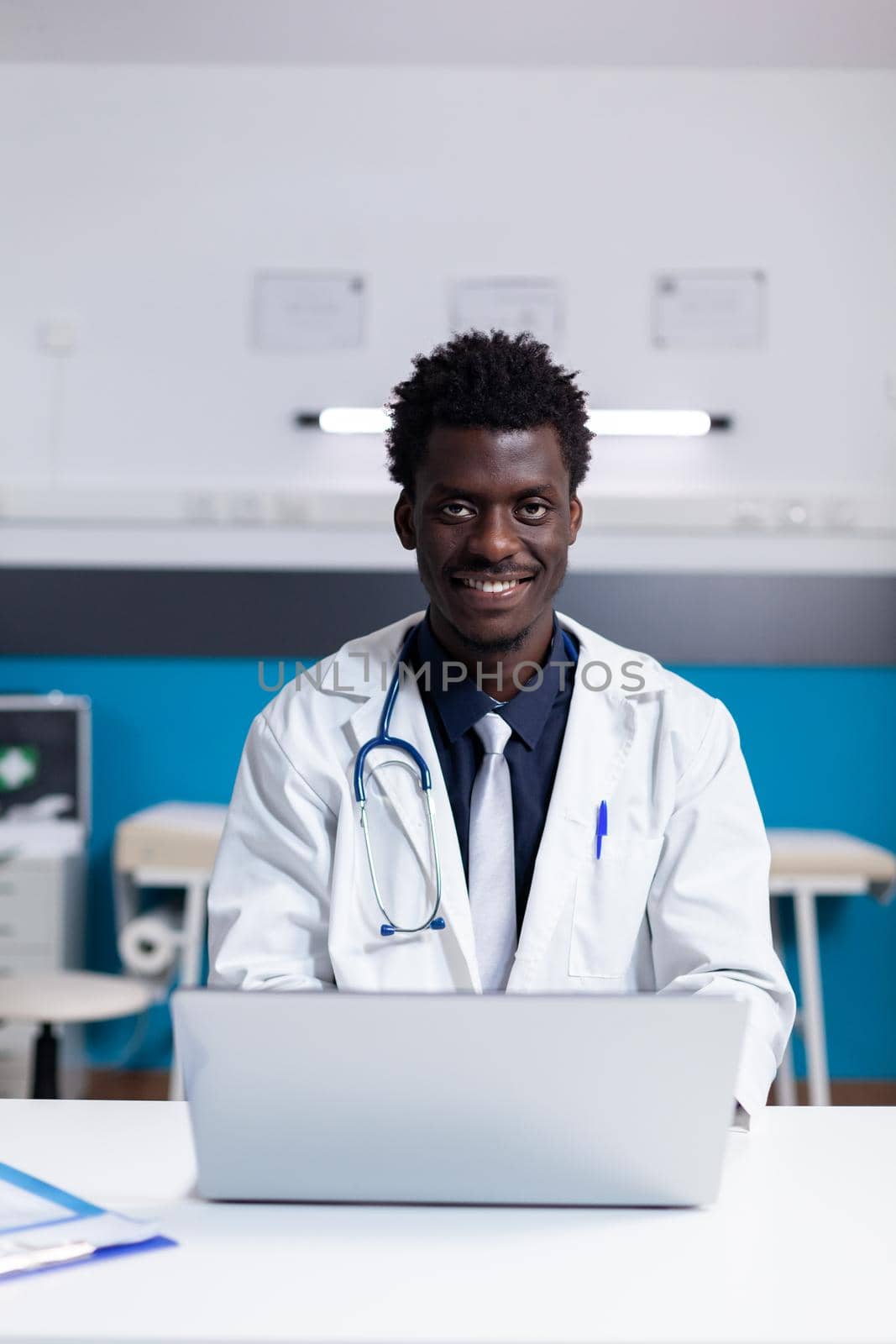 Portrait of african american doctor sitting at desk in healthcare cabinet. Black medic smiling looking at camera while using laptop computer. Person with uniform and stethoscope at clinic