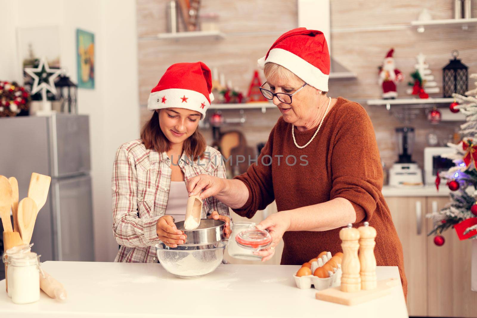 Mature woman and child on christmas day making cookies dough for festive celebration. Happy cheerful joyfull teenage girl helping senior woman preparing sweet cookies to celebrate winter holidays wearing santa hat.